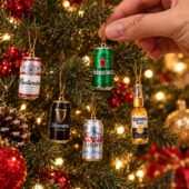 A hand hangs a variety of miniature beer can and bottle Christmas tree ornaments.