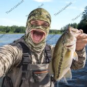 Angler wearing a bass fish mouth face mask holding a freshly caught largemouth bass by the lake.