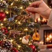 A hand hanging a T-Rex dinosaur ornament on a Christmas tree with other prehistoric figures.