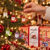 A hand hanging miniature Monin syrup bottle ornaments on a decorated Christmas tree with an advent calendar.